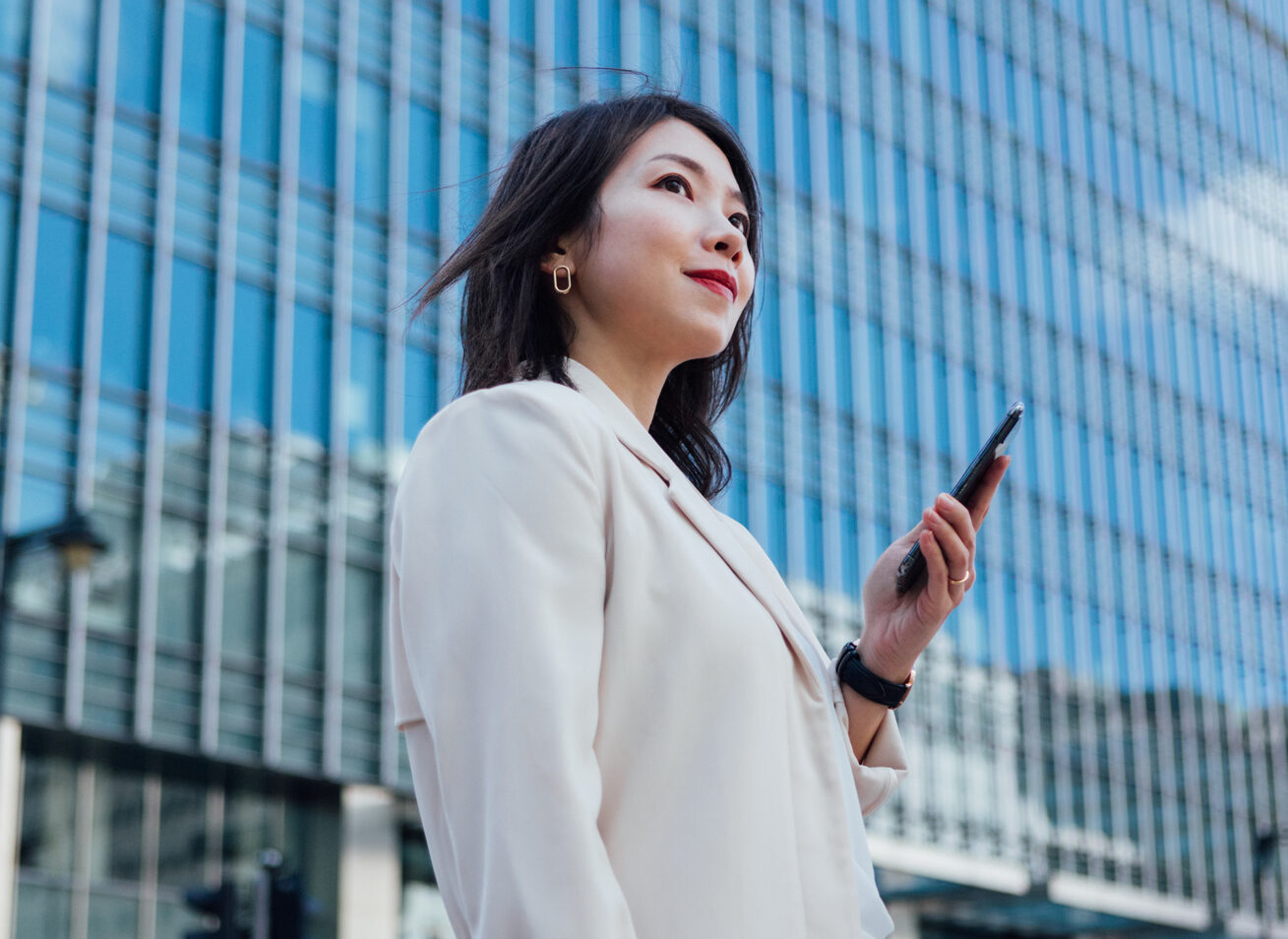 Low angle view of an Asian businesswoman looking away, holding a smart phone, standing against modern buildings in the city. Business on the go concept.