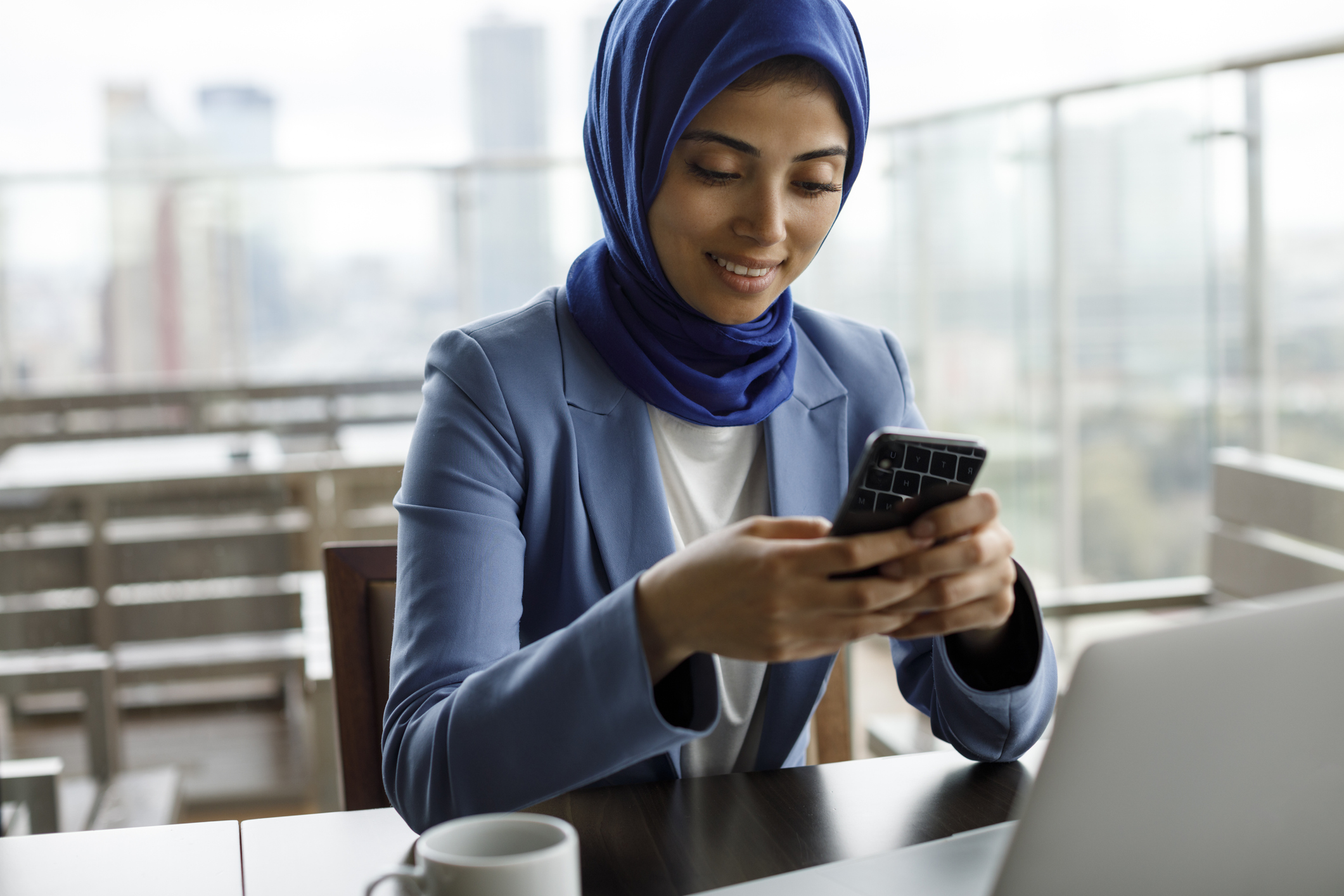 Smiling young muslim woman using mobile phone and laptop at a cafe