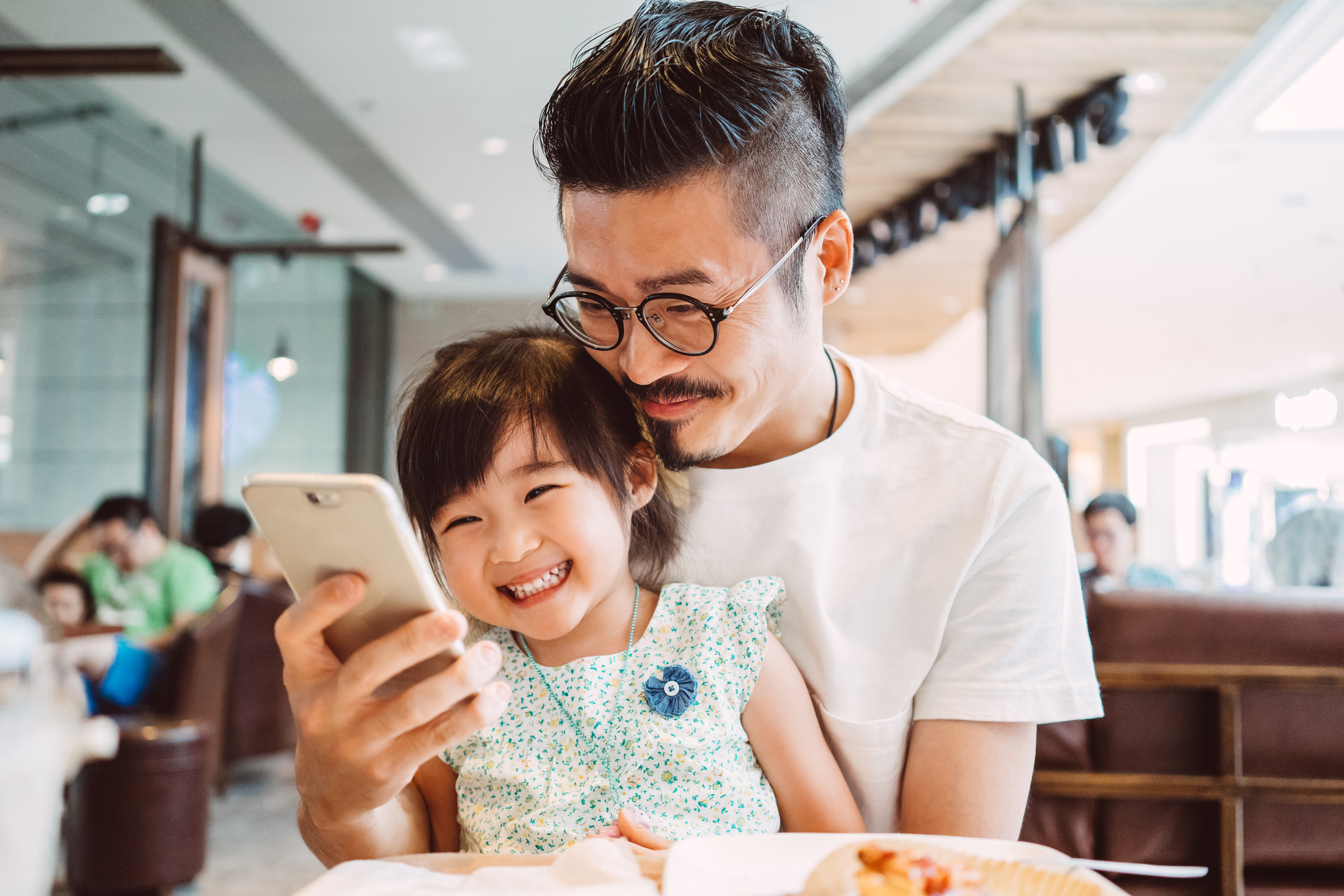 Young dad using smartphone with his little daughter while little daughter sitting on his lap in a coffee shop joyfully